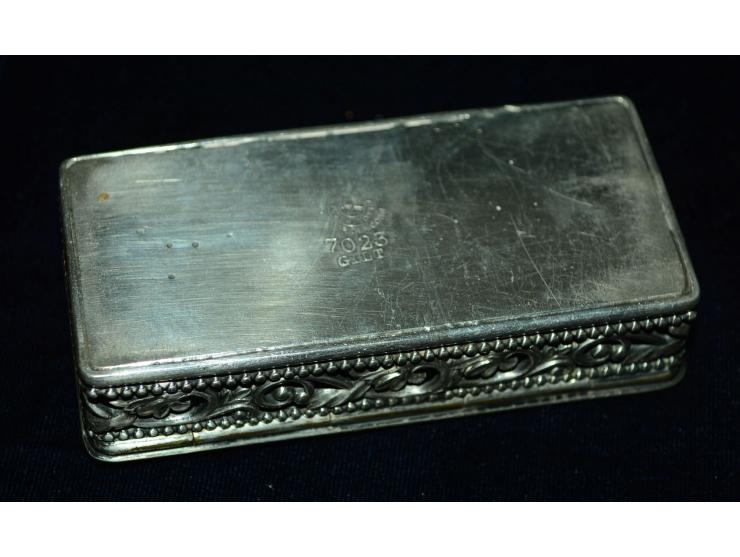 Briefmarkendose aus versilbertem Metal mit getriebenem Bild einer Briefmarke und aufgesetzter Schreibfeder. Die Dose hat zwei
